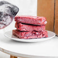Dog looking at a plate of raw meat next to an open box on a small table.